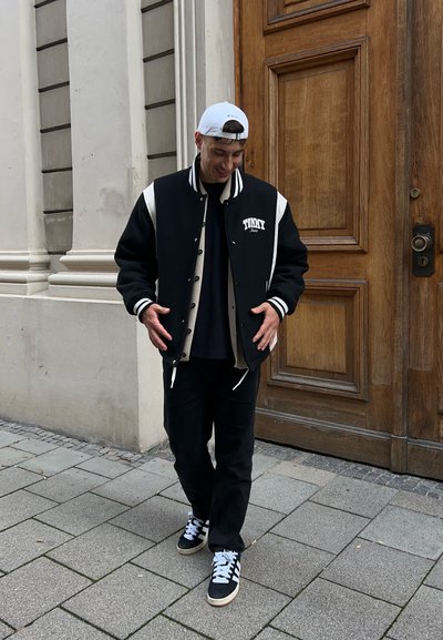 Chaqueta varsity en blanco y negro con acentos laterales, botones en la parte frontal, usada sobre una camiseta negra y combinada con pantalones negros y zapatillas.