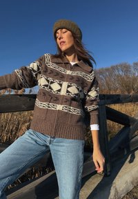 Femme portant un pull marron à motifs, un bonnet tricoté olive et un jean bleu clair, appuyée contre une clôture en bois sous un ciel bleu clair.