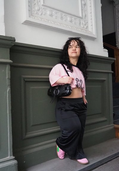 Pantalones de pierna ancha negros, una camiseta corta rosa con texto negro y zapatillas rosas con detalles verdes. Un pequeño bolso negro añade un toque de detalle.
