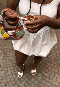 Donna in abito bianco e sandali tiene una borsa di paillettes colorata e una coppa di gelato al cioccolato con un cucchiaio su un pavimento in ciottoli.