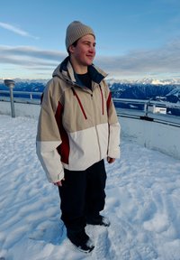 Joven con chaqueta beige y blanca y gorro de lana está en un mirador montañoso nevado con picos cubiertos de nieve a lo lejos bajo un cielo despejado.