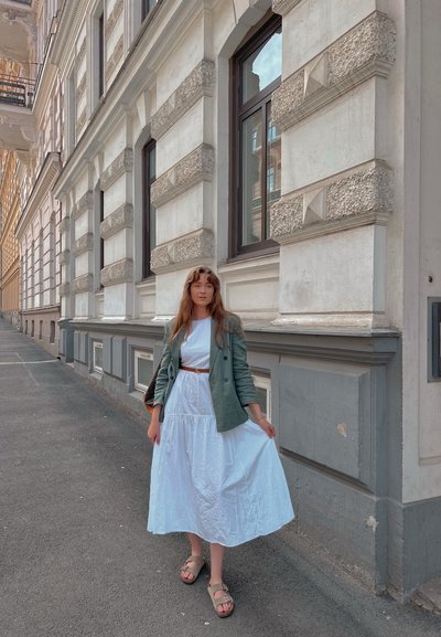 Blazer de lino verde sobre un vestido blanco texturizado hasta la rodilla; sandalias de color beige; entorno urbano con arquitectura histórica y ventanas modernas.