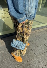 Farmernadrágot és bornyakú csizmát viselő személy kezében tartott, steppelt mintázatú farmer táska, arany lánc pánttal és kör alakú záróelemmel.