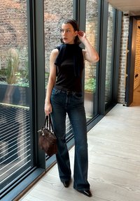 Haut noir sans manches, jeans évasés bleu foncé, sac à main en daim marron, portant un serre-tête, debout dans un espace avec de grandes fenêtres et de la verdure.