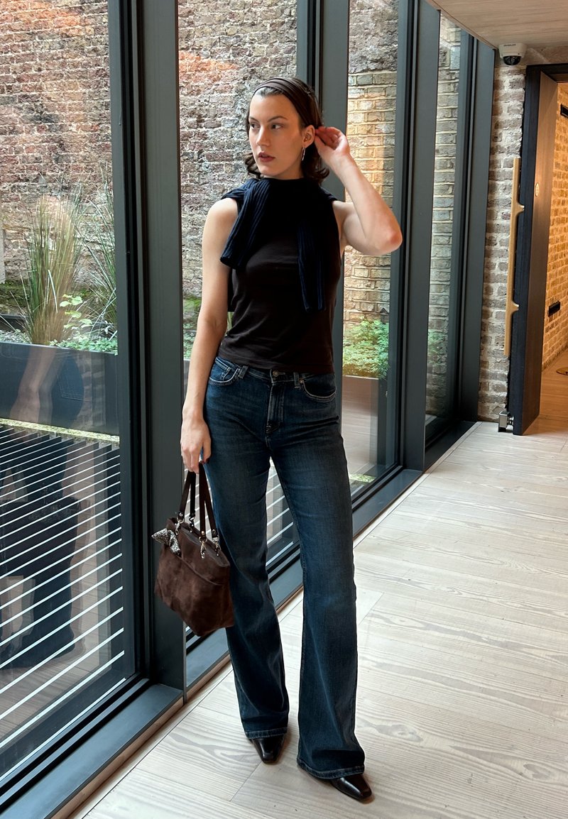 Haut noir sans manches, jeans évasés bleu foncé, sac à main en daim marron, portant un serre-tête, debout dans un espace avec de grandes fenêtres et de la verdure.