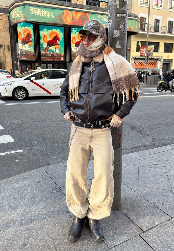 Άνδρας με δερμάτινο μπουφάν, μπεζ παντελόνι, καρό κασκόλ τυλιγμένο γύρω από το κεφάλι και τον λαιμό, γυαλιά ηλίου και καπέλο, στέκεται σε αστικό πεζοδρόμιο κοντά σε φωτεινό σηματοδότη.