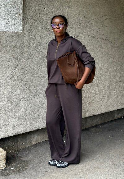Mujer vestida con chándal morado oscuro y zapatillas deportivas, sosteniendo un gran bolso tote de ante marrón, de pie frente a una pared texturizada al aire libre.