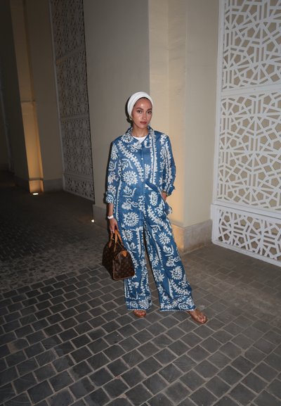 Conjunto de dos piezas azul estampado con diseños florales, pantalones de pierna ancha y camisa abotonada. Los accesorios incluyen un bolso marrón y un pañuelo blanco.
