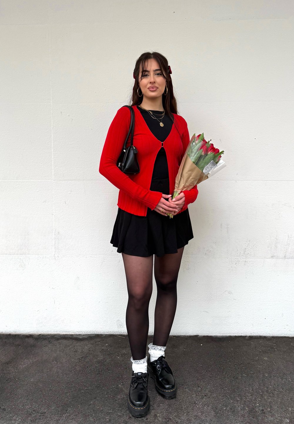 Jeune femme en cardigan rouge et jupe noire tenant un bouquet de tulipes rouges, debout contre un mur blanc.