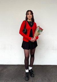 Jeune femme en cardigan rouge et jupe noire tenant un bouquet de tulipes rouges, debout contre un mur blanc.