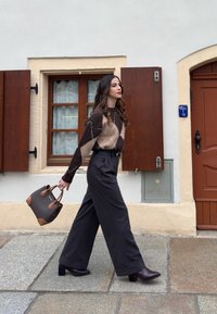 Femme marchant sur un pavement en pierre, vêtue d'un pull marron à motif, d'un pantalon large noir, de bottines à talons, et portant un sac à main marron près d'un bâtiment blanc.