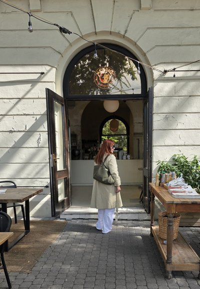 Puerta de madera abierta a un café con una lámpara colgante blanca redonda. Las características exteriores incluyen una pared de piedra beige, un camino de adoquines y muebles de madera.