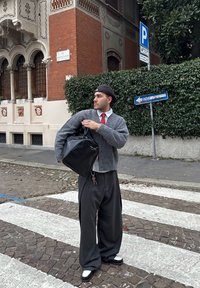 Giovane uomo con cardigan grigio, cravatta rossa e berretto che porta una borsa nera, in piedi su un attraversamento pedonale di ciottoli in una città europea.