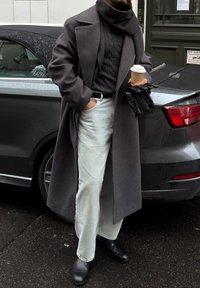 Sivi volna plašč, nošen čez črni pulover s kabelskim vzorcem, kombiniran s svetlo modrimi kavbojkami in črnimi usnjenimi čevlji. V roki drži skodelico kave in rokavice.