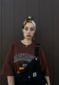 Καφέ μπλουζάκι Carhartt με έντονο λευκό λογότυπο, μαύρες σαλοπέτες με χρυσές λεπτομέρειες, λουλουδάτο μαντήλι και διακοσμητικά σκουλαρίκια. Μονοχρωματικό φόντο.