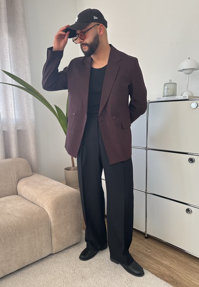 Blazer burdeos oscuro, camiseta negra y pantalones anchos negros. Gorra negra y gafas de sol. La habitación cuenta con muebles claros y vegetación.