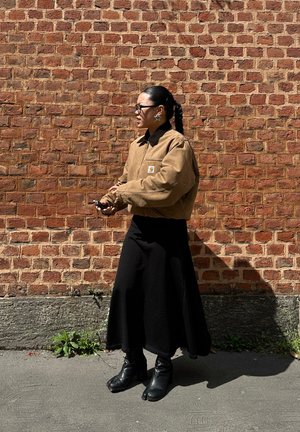 Brown quilted jacket with a black collar, black long skirt, and black heeled boots. Standing against a textured red brick wall.