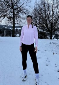 Γυναίκα με ροζ μακρυμάνικη μπλούζα, μαύρα κολάν και λευκά αθλητικά παπούτσια, στέκεται σε χιονισμένο έδαφος με φυλλοβόλα δέντρα στο φόντο.