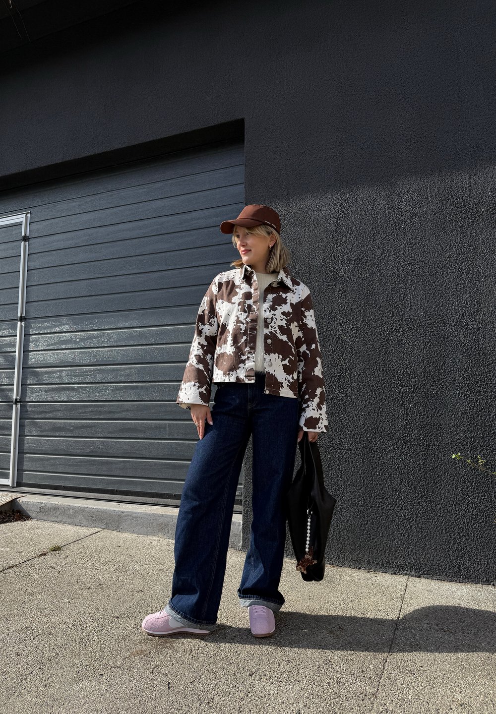 Braune und weiße gemusterte Jacke, hellgraues Oberteil, dunkelblaue weite Jeans, hellrosa und graue Schuhe und eine dunkle Tasche. Braune Mütze auf dem Kopf.