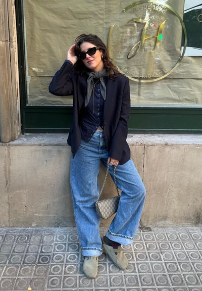 Mujer con gafas de sol, bufanda, blazer oscuro, camisa abotonada, vaqueros anchos y zuecos beige, de pie contra una pared sosteniendo un bolso de cadena.