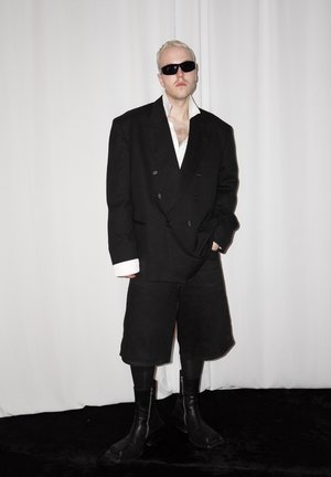 Hombre con cabello corto rubio y gafas de sol lleva una americana negra extragrande, pantalones cortos negros largos, botas negras y camisa blanca, de pie frente a un fondo blanco.