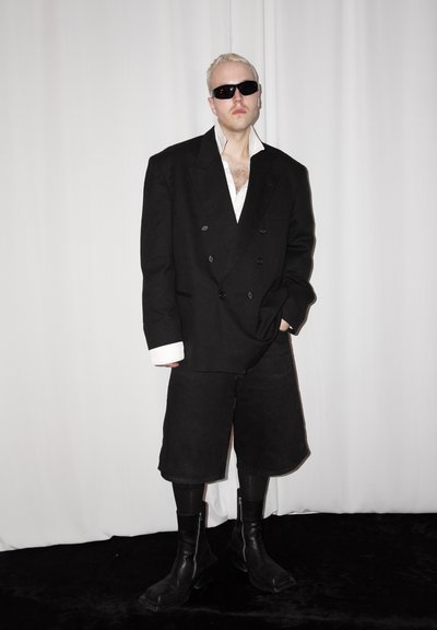Hombre con cabello corto rubio y gafas de sol lleva una americana negra extragrande, pantalones cortos negros largos, botas negras y camisa blanca, de pie frente a un fondo blanco.