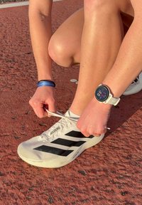 Personne portant une montre connectée blanche qui fait ses lacets sur une chaussure de course blanche Adidas Adizero sur une piste extérieure rouge.