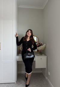 Femme en robe noire et talons tenant un sac à main noir, appuyée contre une armoire blanche près d'une coiffeuse avec un bouquet de fleurs.