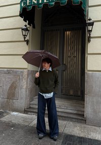 Jeune personne debout sous un auvent vert, tenant un parapluie ouvert, vêtue d'un pull foncé, d'une chemise rayée et d'un pantalon large sur le trottoir de la ville.