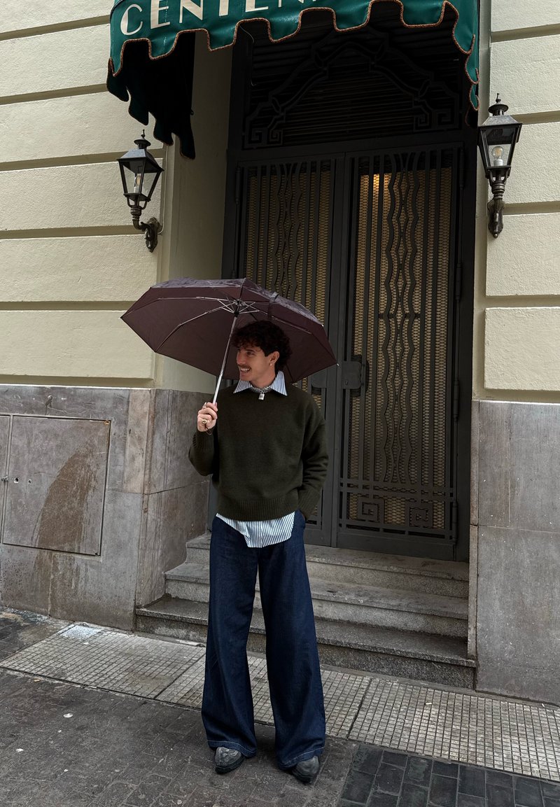 Giovane in piedi sotto una tenda verde, che tiene aperto un ombrello, indossa un maglione scuro, una camicia a righe e pantaloni larghi sul marciapiede della città.