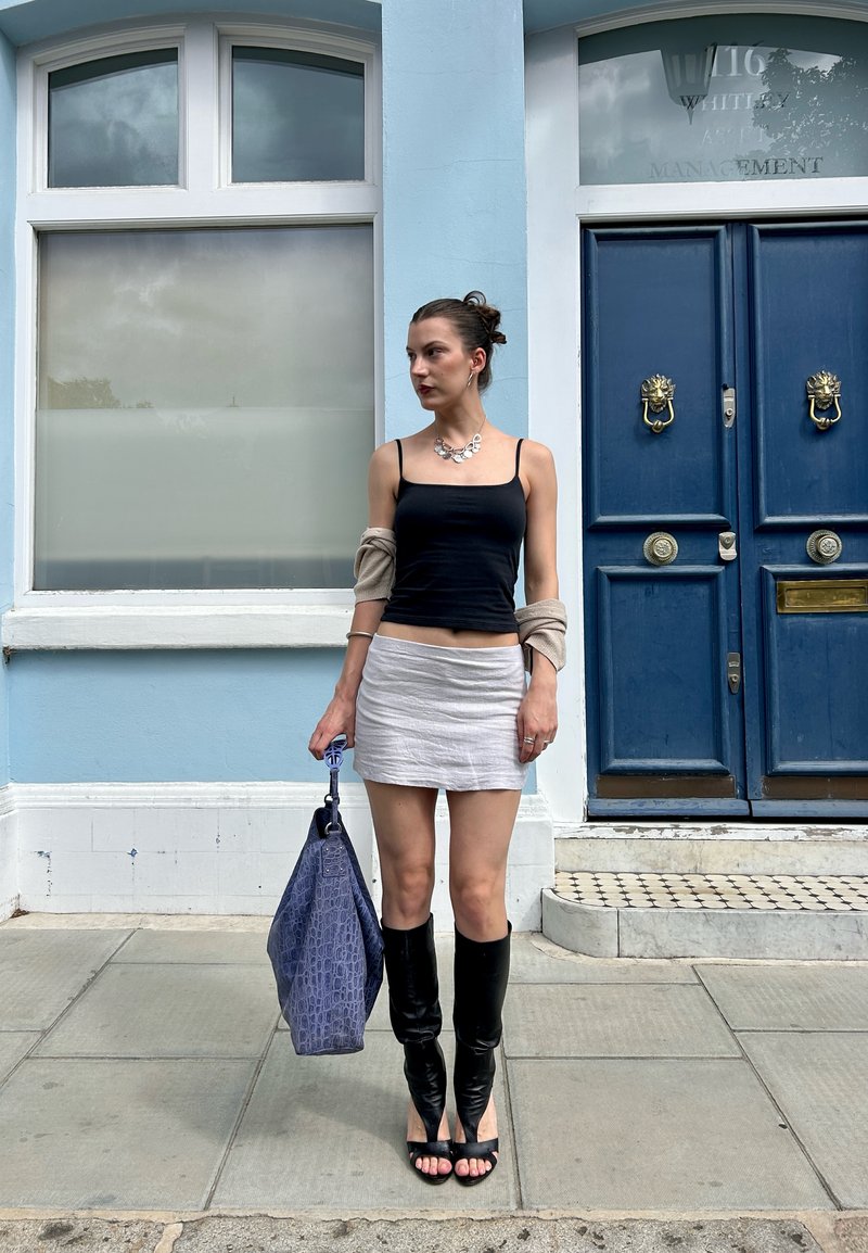 Woman standing on sidewalk wearing black tank top, white mini skirt, black open-toe boots, holding large blue bag, in front of blue wall and door.