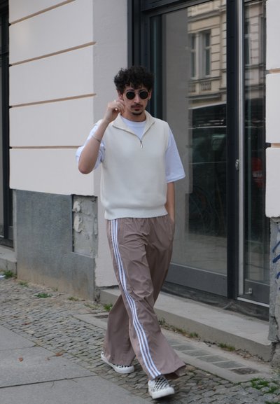 Suéter sin mangas blanco sobre una camiseta blanca, combinado con pantalones deportivos marrón claro con franjas laterales blancas y zapatos con un patrón en blanco y negro.