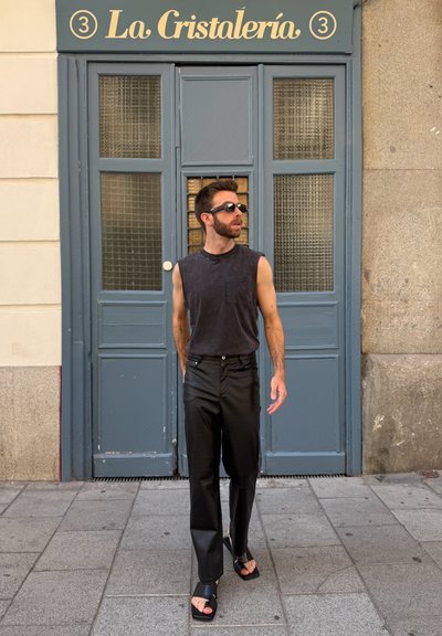 Top negro sin mangas, pantalones negros con un ligero brillo y sandalias negras tipo slide. De pie frente a una puerta doble azul con paneles de vidrio.