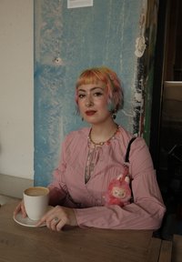 Una persona con cabello en tonos pastel lleva una blusa rosa de manga larga. Sostiene una taza blanca sobre un platillo, con un llavero de felpa rosa en su bolso.