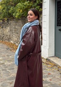 Femme avec une longue tresse portant un manteau en cuir bordeaux et une écharpe bleu clair, debout dans une rue pavée près d'une porte verte et d'un mur en pierre.