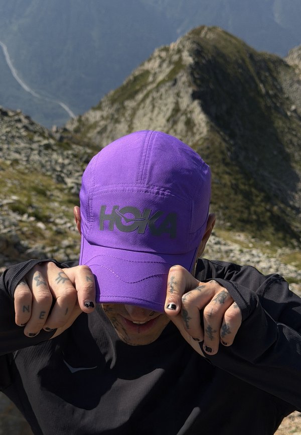 Person with tattooed hands adjusting a purple HOKA cap, wearing black clothing, standing outdoors with rocky mountains in the background.