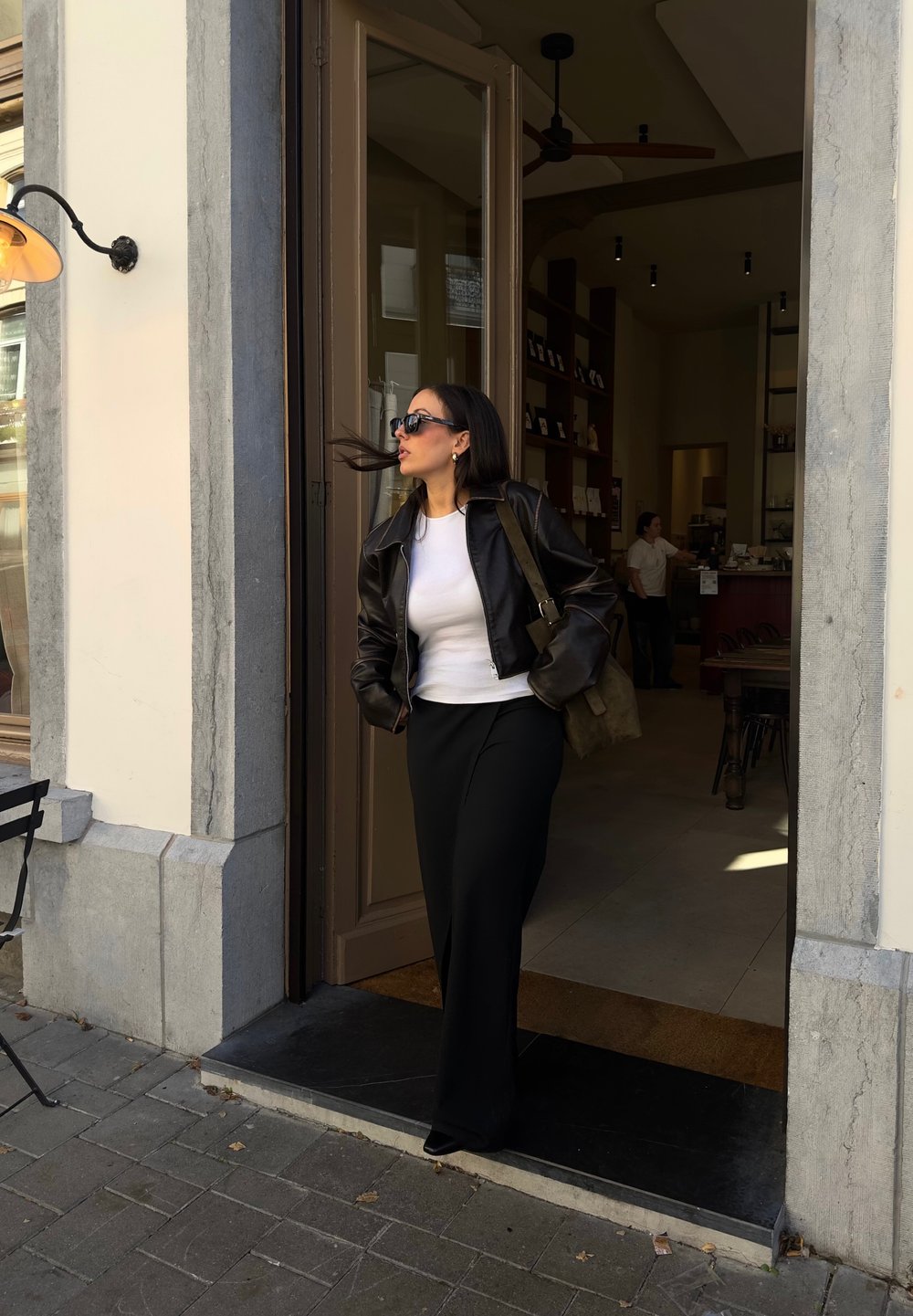 Μαύρο δερμάτινο μπουφάν, λευκή μπλούζα και μακριά μαύρη φούστα. Μαύρα γυαλιά ηλίου και τσάντα ώμου. Σταθείς σε μια πόρτα με πέτρινο δάπεδο.