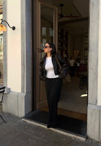 Μαύρο δερμάτινο μπουφάν, λευκή μπλούζα και μακριά μαύρη φούστα. Μαύρα γυαλιά ηλίου και τσάντα ώμου. Σταθείς σε μια πόρτα με πέτρινο δάπεδο.