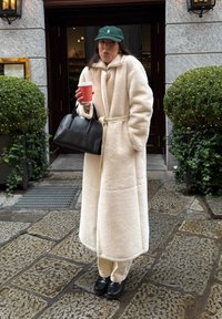 Krém színű, bolyhos hosszú kabát övvel, bézs nadrággal és fekete cipővel párosítva. Vörös kávésbögrét és fekete kézitáskát tart. Zöld sapka.