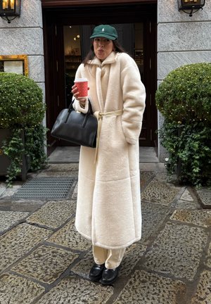 Abrigo largo y esponjoso de color crema con cinturón, combinado con pantalones beige y zapatos negros. Sostiene una taza de café roja y un bolso negro. Lleva una gorra verde.