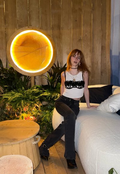 Joven mujer con cabello rojo, vestida con una blusa blanca y jeans negros, sentada en un sofá blanco junto a una decoración circular de pared de madera iluminada y plantas.