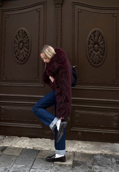 Abrigo de piel sintética burdeos, jeans de mezclilla azul oscuro con los puños enrollados, mocasines negros y calcetines blancos. El fondo presenta una puerta marrón ornamentada.