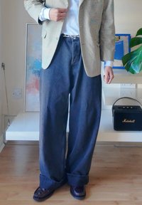 Wide-leg dark denim jeans paired with a light patterned blazer and white shirt; shiny brown loafers complete the outfit.