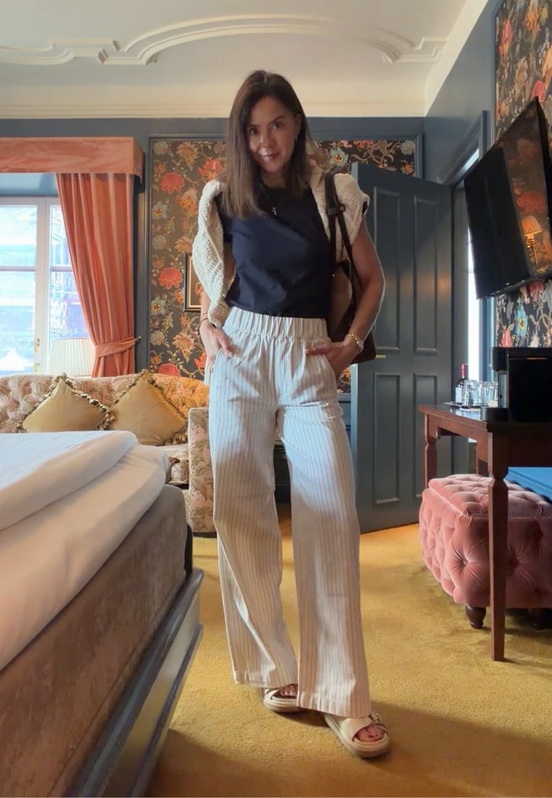 Femme debout dans une pièce de style vintage portant un top noir, un pantalon large blanc à rayures, des sandales et un pull blanc drapé sur ses épaules.