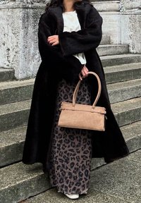 Lang svart fuskepelskåpe over et beige skjørt med leopardmønster, kombinert med en liten beige håndveske og hvit topp, stående på steintrapper.