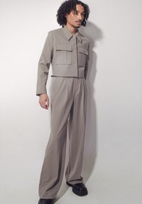 Veste courte gris clair avec deux poches avant et un pantalon taille haute assorti. Tissu texturé, coupe ajustée, associée à des chaussures noires.