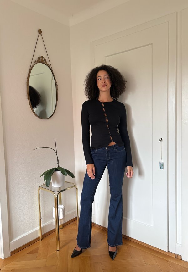 Femme aux cheveux bouclés portant un haut noir à manches longues et un jean bleu foncé, se tenant près d'une porte blanche et d'une petite table avec une plante en pot et un miroir.