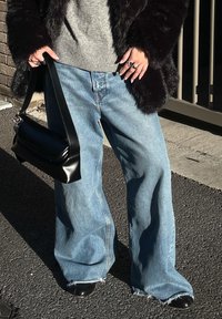 Personne portant un jean large bleu, des chaussures pointues noires, un pull gris, un manteau en fourrure noir, tenant un sac à main en cuir noir à l'extérieur au soleil.