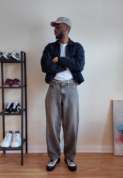 Hombre con gorra, chaqueta oscura, camisa blanca, vaqueros grises holgados y zapatillas está con los brazos cruzados junto a un zapatero y una pintura abstracta.