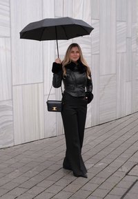 Veste en cuir noire avec accents en fourrure, pantalons noirs taille haute, et un sac à main noir. Tenant un parapluie noir contre un mur en pierre claire.